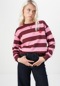 Prajčasta, maroon in roza majica s kapuco s zaobljenim izrezom in izvezenim rdečim srčastim detajlom. Kombinirana z temno modrimi kavbojkami.