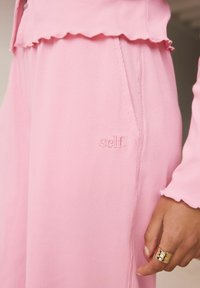 Ružové ribované nohavice z látky so bočnou kapsou, s vyšitým logom "self." Látka má mäkkú textúru a splývavý strih.