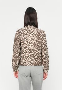 Personne aux cheveux foncés mi-longs portant un haut marron et beige à manches longues avec un imprimé léopard et un pantalon gris clair, vue de dos.