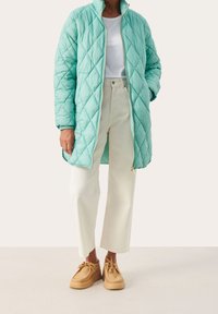 Gequiltede mintgroene lange jas met hoge kraag, ritssluiting en zijzakken, gecombineerd met witte wijde broekspijpen en tan schoenen.