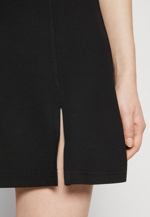 Close-up van de zoom van een zwarte rok met een voorsplit, met een hand met een lichte huidtoneel die naast de rok rust.