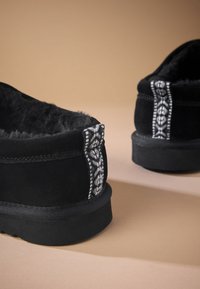 Chaussures noires doublées en polaire avec une tige en suède doux. Présentent des coutures décoratives argentées et blanches le long du talon. Semelle en caoutchouc noire plate.