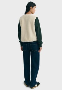 Kvinde vendt væk iført en cremefarvet ærmeløs fleecevest, mørkegrøn langærmet skjorte, brede blå jeans og sorte sneakers med beige såler.
