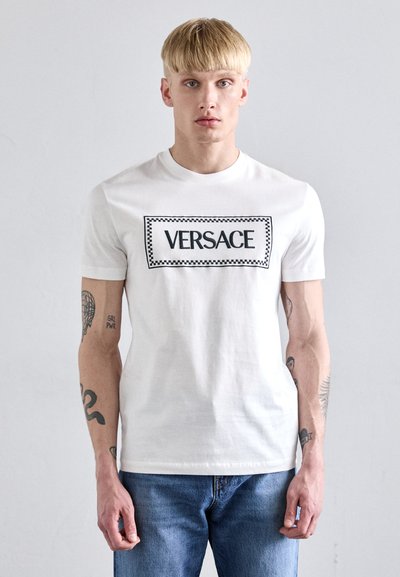 Camiseta de algodón blanca con un gran logotipo rectangular negro "VERSACE" centrado en la parte delantera, mangas cortas y un cuello redondo estándar.