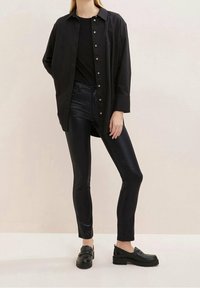 Schwarze Hemdbluse über einem schwarzen Top, kombiniert mit glänzenden schwarzen Leggings und schwarzen Loafers. Glatte Texturen und geschichtete Designdetails.