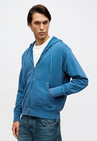 Νέος άνδρας με καστανά μαλλιά, φορώντας μπλε φούτερ με φερμουάρ, λευκό πουκάμισο και μπλε τζιν, χέρι στην τσέπη, κοιτάζοντας μπροστά με ουδέτερη έκφραση.