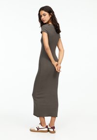Figurbetontes, kurzärmeliges Midikleid aus dunkelolivgrünem Stoff. Das Model trägt weiße Sneakers mit burgunderfarbenen Akzenten und beigen Sohlen.