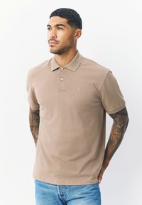 Beżowy polo z kołnierzykiem, trzema guzikami na plastrze i małym haftowanym logo. Posiada krótkie rękawy i gładką teksturę.