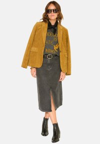 Blazer en velours côtelé jaune sur un sweat-shirt noir à motif, jupe en denim noir avec une fente, et bottines noires. Accessoires comprenant des lunettes de soleil.