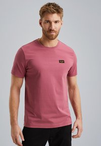 T-shirt in cotone rosa con collo rotondo, maniche corte e una piccola etichetta logo nera sul lato sinistro del petto. Texture liscia, design aderente.