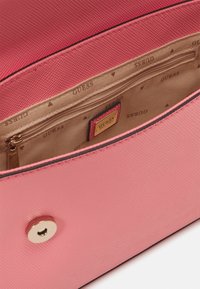 Różowa torebka o fakturze z beżowym wnętrzem, kieszonką na zamek błyskawiczny, zapięciem magnetycznym oraz logo marki Guess na etykiecie wewnętrznej.