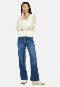 Maglione crema a maglia con maniche in pizzo trasparente, abbinato a jeans in denim a gamba larga blu chiaro e sneakers beige. Design semplice e a strati.
