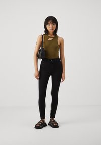 Jauna moteris stovi, vilkėdama žalią be rankovių palaidinę, juodas siauras kelnes, juodas šlepetes ir nešdama mažą juodą petinę kuprinę.