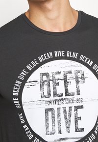 Camiseta de algodón gris oscuro con un diseño gráfico redondo que presenta el texto "DEEP DIVE" en una fuente blanca desgastada y acentos circulares.