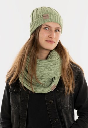 Groene gebreide beanie met een ribbelpatroon en logo-label, gecombineerd met een chunky gebreide infinity-sjaal in dezelfde kleur. Zwarte spijkerjack.