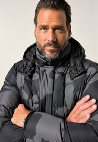 Uomo con barba e capelli corti che indossa un giubbotto imbottito nero e grigio con cappuccio, braccia incrociate, che guarda dritto davanti a sé su uno sfondo semplice.