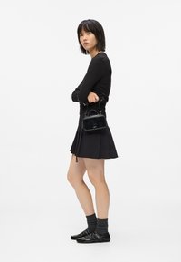 Kobieta z prostymi czarnymi włosami, ubrana w czarną bluzk�ę z długim rękawem, plisowaną czarną spódnicę, szare skarpetki, czarne buty, trzymająca małą czarną torebkę.