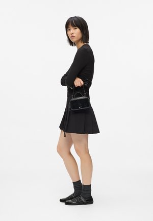 Fekete egyenes hajú nő fekete hosszú ujjú felsőt, rakott fekete szoknyát, szürke zoknit és fekete cipőt visel, miközben egy kis fekete táska van a kezében.