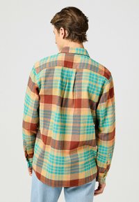 Camisa de franela en cuadros amarillos, azules y marrones. Cuenta con un cuello estándar, puños abotonados y un canesú en la espalda con un detalle de pliegue.