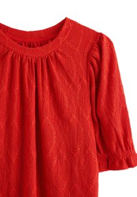 Rød tekstureret stofkjole med øjehulsmønstre i diamantform, rynket halsudskæring og lange, puffede ærmer med elastiske manchetter.