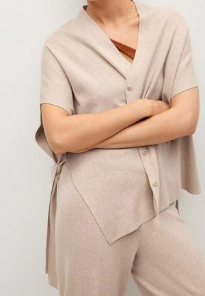 Person trägt beige kurzärmelige, geknöpfte Strickjacke über passenden Hosen, mit verschränkten Armen und darunter ein rostfarbenes Top sichtbar.