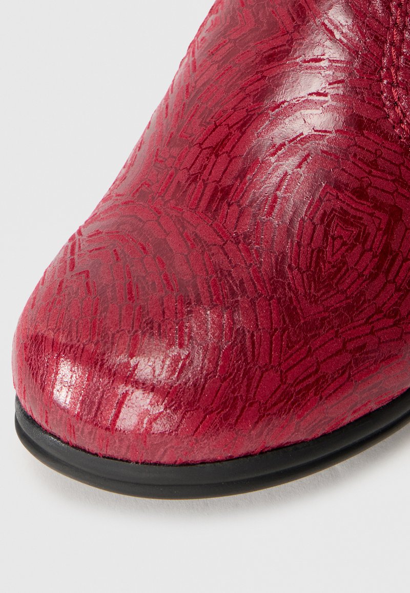 Scarpa rossa dal finish lucido, con una texture in rilievo e un motivo geometrico sulla parte superiore e punta rotonda. Suola nera visibile sotto.