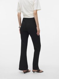 Pantaloni negri evazați cu un model texturat, talie înaltă și croială dreptă. Modelul poartă un tricou alb simplu, scurt și sandale cu toc mic.