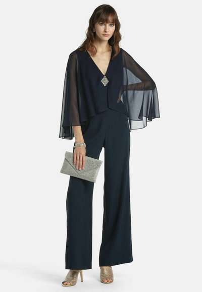 Marineblauer Overall mit durchsichtigen Cape-Ärmeln, tiefem V-Ausschnitt, silberner Broschenakzentuierung, kombiniert mit einer silbernen Clutch und goldenen offenen Sandaletten.