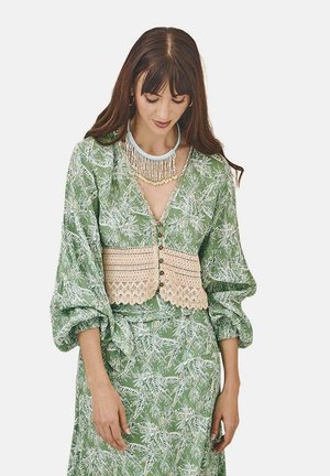 Blusa verde con estampado y mangas abullonadas, con una superposición de ganchillo beige y botones, combinada con una falda a juego.