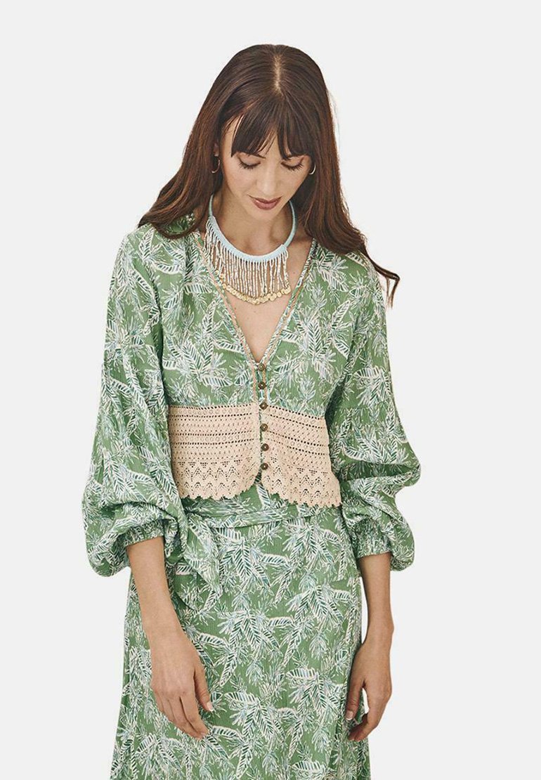 Blusa verde con estampado y mangas abullonadas, con una superposición de ganchillo beige y botones, combinada con una falda a juego.