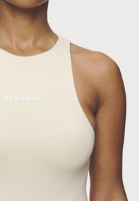 Body senza maniche beige con collo alto e texture leggera, con logo bianco "PEGADOR" sul petto.