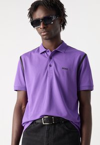 Μωβ πόλο μπλουζάκι με κλασικό γιακά, κοντά μανίκια και μαύρες λεπτομέρειες στους ώμους; διαθέτει ένα μικρό μαύρο λογότυπο στο στήθος.