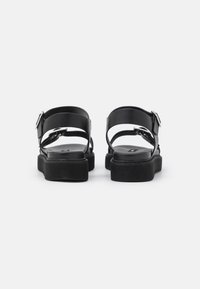 Černé sandály s plochou podrážkou, dvojitými popruhy kolem kotníku a nastavitelnou kovovou sponou. Hladký kožený povrch a minimalistický design.