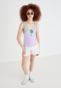 Lilla ribbet tanktop med grønn kant og sløyfedetalj, paret med hvite shorts med bølgete kant. Modellen har på seg blå solbriller og mintgrønne sneakers.
