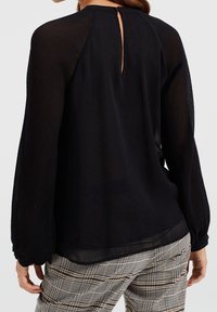 Zwarte blouse met sheer gestreepte mouwen, een keyhole detail op de rug en een ontspannen pasvorm, gecombineerd met een broek in een ruitpatroon.