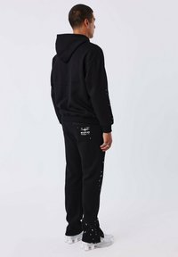 Zwarte hoodie en broek met witte verfspatten. De stof is zacht en heeft een ontspannen pasvorm. De broek heeft een logopatch en witte accenten aan de zijkant.
