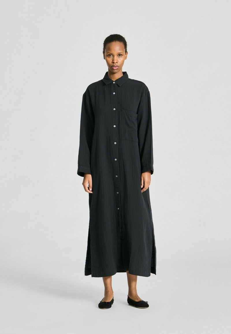 Femme debout portant une longue robe-chemise noire boutonnée avec col et fentes latérales, chaussant des ballerines noires sur un fond clair uni.