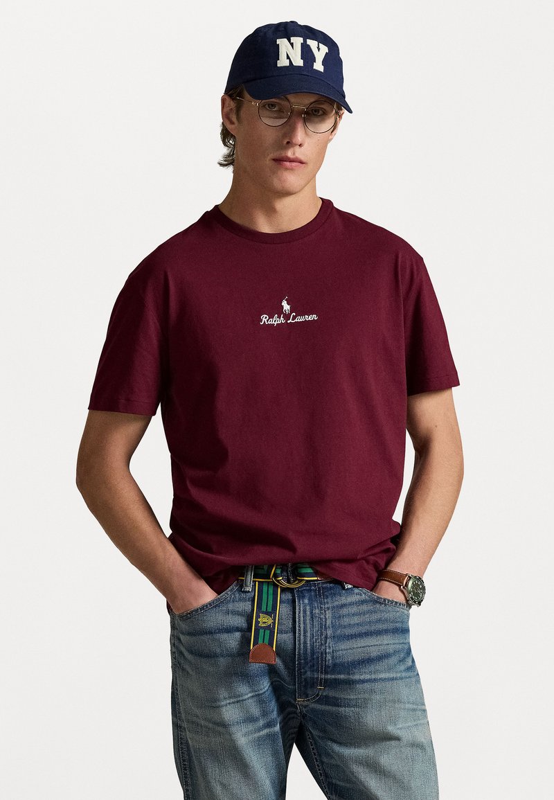 Camiseta de algodón burdeos con el logo bordado de "Ralph Lauren", combinada con vaqueros azules y un cinturón de rayas multicolores. Los accesorios incluyen gafas y una gorra azul marino.