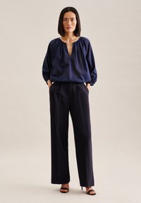 Marineblaue Bluse mit lockerer Passform und gerafftem Ausschnitt, kombiniert mit hochgeschnittenen, weiten Marinehosen und schwarzen High-Heel-Sandalen.