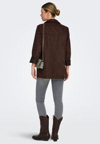 Bruine oversized soepel suède shirtjack met opgerolde mouwen, detail op achterkant, gecombineerd met grijze skinny jeans en bruine cowboylaarzen. Zilveren kettingtas.