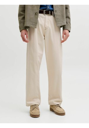 Férfi bézs színű, bő szárú nadrágban, barna övvel, világosbarna mokaszin jellegű cipőben, kék ingben és szürke dzsekiben, fehér háttér előtt áll.