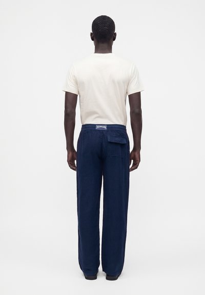 Homme debout de dos, portant une chemise blanche à manches courtes et un pantalon ample bleu foncé avec une poche arrière et une étiquette à la taille.