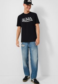 Juodas medvilninis marškinėliai su baltu "Von Dutch" logotipu, derinami su mėlynomis džinsinėmis kelnėmis su aplikacijomis ir juodais sportbačiais su žaliais padu.