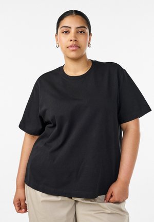 Kobieta z ciemnymi włosami upiętymi w niskiego koka ma na sobie czarny t-shirt z krótkim rękawem i beżowe spodnie, stojąc na tle gładkiego białego tła.