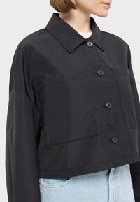 Jachetă neagră scurtă, realizată dintr-un material ușor, cu închidere prin nasturi, două buzunare pe piept și guler. Textură netedă, cu un design minimalist.
