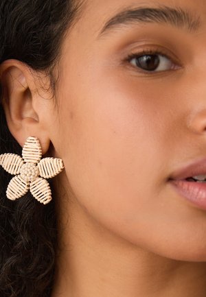Close-up of a person wearing a large beige flower-shaped earring with braided and beaded details on the left ear.