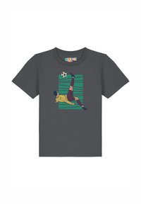 Grijze t-shirt met een afbeelding van een voetballer in een gele trui die een trap uitvoert, tegen een achtergrond van groene horizontale strepen.