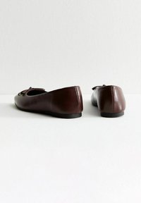 Un paio di ballerine in pelle marrone scuro con piccoli fiocchi, viste da dietro su una superficie bianca e contro uno sfondo bianco.
