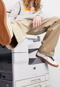 Vita träningsskor med marinblå snören och gula sulor, beige byxor med lös passform och en ljusgrå huvtröja med en orange Nike-logotyp, sittande på en skrivare.