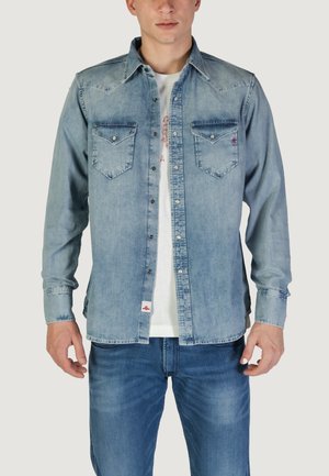 Camicia di jeans azzurro chiaro con due taschini sul petto, colletto a punta e bottoni metallici. Indossata sopra una t-shirt bianca e abbinata a jeans scuri.